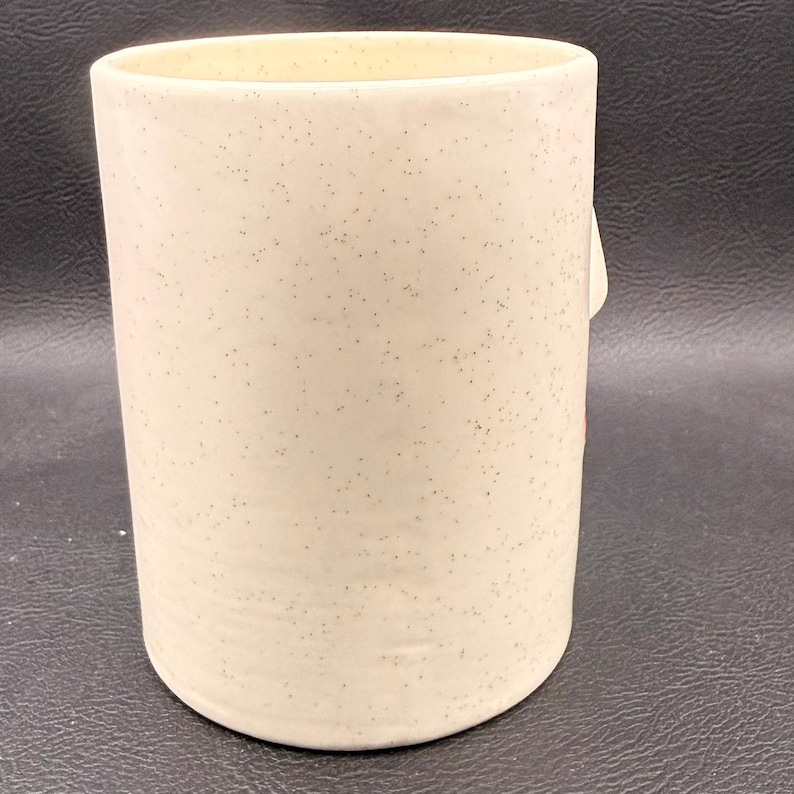 May include: A cylindrical ceramic mug with a speckled, cream-coloured finish. The mug has a smooth, rounded base and a straight, slightly flared top. The exterior is covered in small, dark speckles, giving it a rustic appearance. The mug is empty.