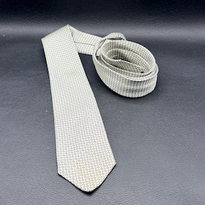 May include: A necktie featuring a light beige fabric base with a repeating pattern of small, dark brown geometric shapes. The tie is laid out flat with the bottom portion extended and the top section coiled into a circular shape on the surface.