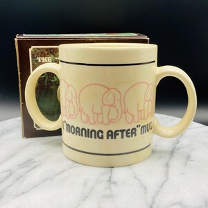 May include: A cream-colored ceramic mug with two handles. The mug has a black line around the top and bottom and features a line of pink elephants with the text "The Morning After" mug.
