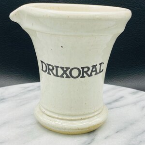 May include: Cream-colored ceramic pitcher with a flared top and a small spout. The word "DRIXORAL" is printed in black on the front of the pitcher. The pitcher has a rounded base and a speckled finish.