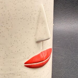 May include: A ceramic mug with a face design. The mug is off-white with small black speckles. A raised nose and red lips are featured on the front of the mug. The mug is a unique and artistic piece.