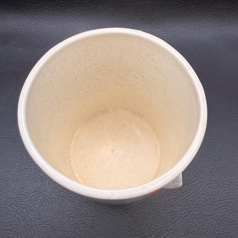 May include: Cream ceramic mug with a speckled interior and a white rim. The mug has a small handle and a red accent near the base. The mug is empty and appears clean.