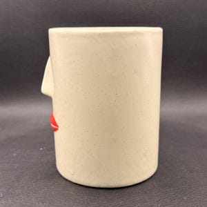 May include: A beige ceramic mug with a face design. The mug has a nose and red lips on the side. The mug is cylindrical and has a speckled texture.