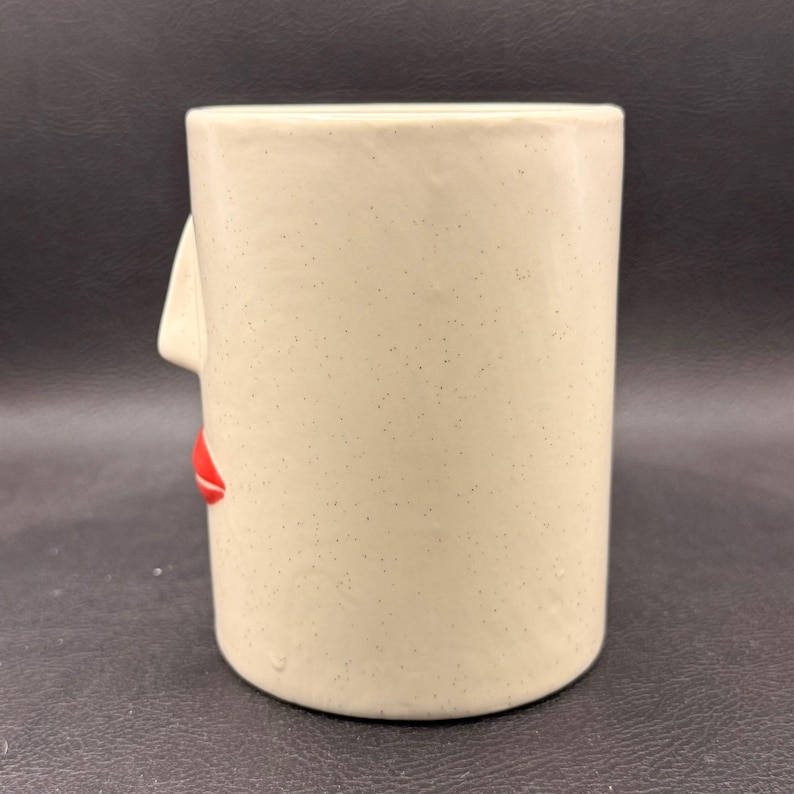May include: A beige ceramic mug with a face design. The mug has a nose and red lips on the side. The mug is cylindrical and has a speckled texture.