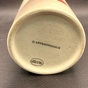 May include: Close-up of the base of a ceramic mug. The base is speckled with small dark spots and features the text "by ANTHROPOLOGIE" and "Made in China Fabriqu&eacute; en Chine". The mug has a cream-colored base.