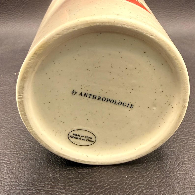 May include: Close-up of the base of a ceramic mug. The base is speckled with small dark spots and features the text "by ANTHROPOLOGIE" and "Made in China Fabriqu&eacute; en Chine". The mug has a cream-colored base.