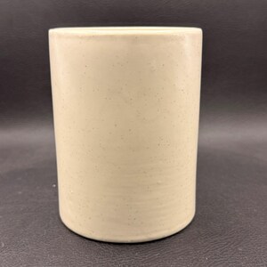 May include: A cylindrical, cream-coloured ceramic container with a speckled surface. The container has a smooth, rounded base and a straight, open top. The neutral colour and simple design suggest a minimalist aesthetic.