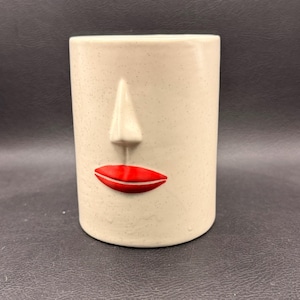 May include: A cylindrical ceramic vase with a face design. The vase is off-white with a textured surface and a raised nose. The lips are painted bright red. The vase is approximately 13 cm tall.