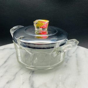 May include: A clear glass sugar bowl with a silver lid and a floral design on the knob. The bowl has a faceted design and two handles.