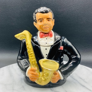 May include: A black and gold ceramic teapot shaped like a man in a tuxedo holding a saxophone. The man has a brown complexion and is wearing a red bow tie.