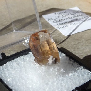 Puede incluir: Un cristal de topacio marr&oacute;n claro con un aspecto transparente y v&iacute;treo. El cristal est&aacute; sentado en una peque&ntilde;a caja de espuma blanca. La etiqueta dice "Topacio Al2(F,OH)2SiO4 Ortorr&oacute;mbico Thomas Range Juab Co."