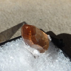 Puede incluir: Un cristal transl&uacute;cido de color marr&oacute;n anaranjado con forma puntiaguda. El cristal est&aacute; sobre una superficie blanca texturizada.