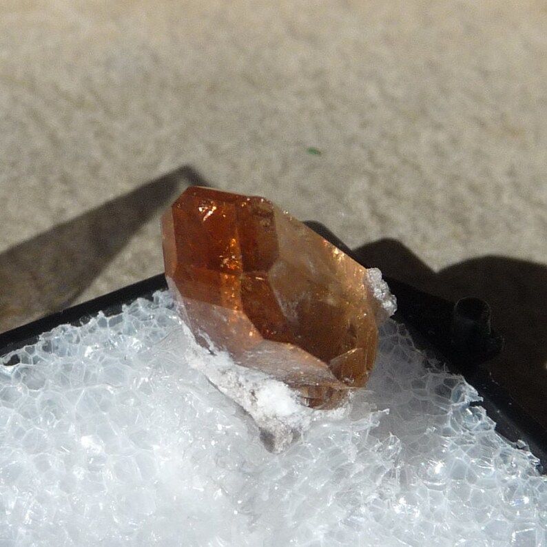 Puede incluir: Un cristal transl&uacute;cido de color marr&oacute;n anaranjado con forma puntiaguda. El cristal est&aacute; sobre una superficie blanca texturizada.