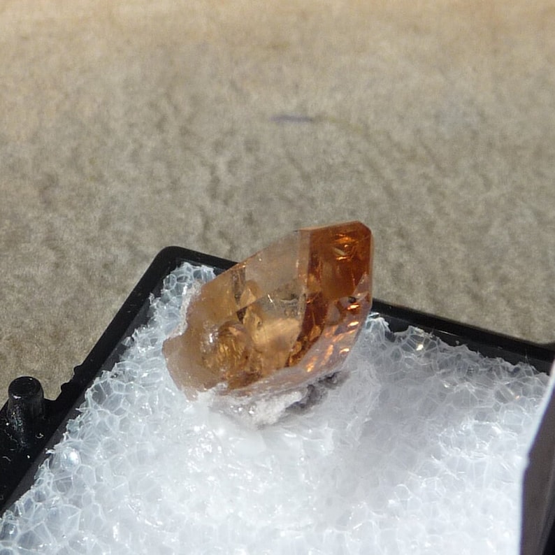 Puede incluir: Un solo cristal de topacio de color naranja-marr&oacute;n claro con una punta puntiaguda. El cristal est&aacute; sentado sobre una superficie de espuma blanca dentro de una caja negra.