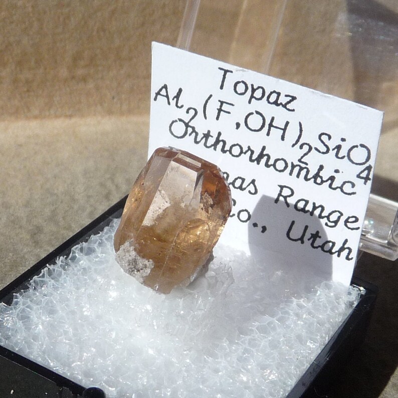 Puede incluir: Un cristal de topacio marr&oacute;n claro con una etiqueta que dice "Topaz Al2(F,OH)2SiO4 Ortorr&oacute;mbico Was Range Co., Utah".