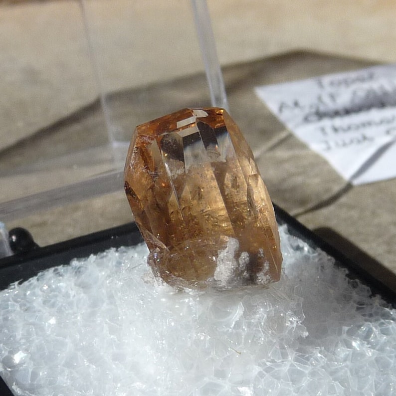 Puede incluir: Un cristal de topacio marr&oacute;n claro y facetado con un ligero tono dorado. El cristal est&aacute; sentado sobre un lecho de hielo picado blanco.