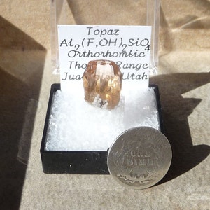 Puede incluir: Un cristal de topacio marr&oacute;n con estr&iacute;as, etiquetado "Topaz Al2(F,OH)2SiO4 Orthorhombic Tho Range Jua Utah" sobre un fondo blanco. Una moneda de plata est&aacute; en el fondo.