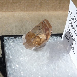 Puede incluir: Un cristal de topacio transparente de color marr&oacute;n claro con una punta puntiaguda. El cristal est&aacute; sentado sobre una superficie blanca de espuma. La etiqueta dice "Al (F) Topaz Thomas Juab Co."