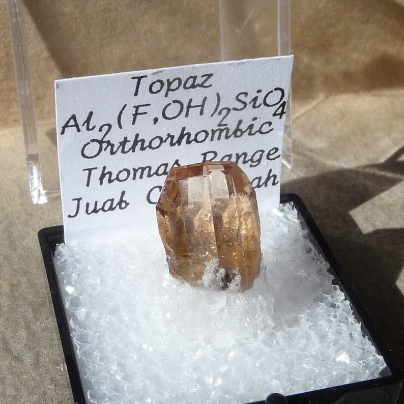 Puede incluir: Un cristal de topacio marr&oacute;n con una superficie clara y facetada. El cristal est&aacute; etiquetado como "Topacio Al2(F,OH)2SiO4 Ortorr&oacute;mbico Thomas Range Juab C ah"