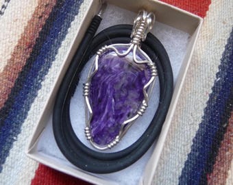 May include: A purple charoite pendant with a silver wire wrap, presented on a black cord. The pendant is inside a small, rectangular box with white padding. The stone has a swirling pattern of purple and white.