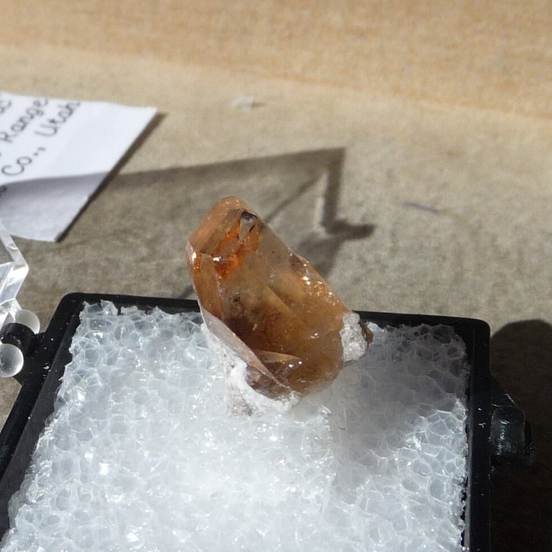 Puede incluir: Un solo cristal transparente de color marr&oacute;n anaranjado con una punta puntiaguda, sentado sobre una superficie blanca de espuma en una caja negra.