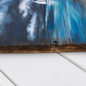 May include: A close-up of a wooden sign with a blue and white painted seascape. The sign has a rustic, weathered look and is perfect for a coastal-themed home decor.