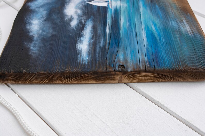 May include: A close-up of a wooden sign with a blue and white painted seascape. The sign has a rustic, weathered look and is perfect for a coastal-themed home decor.
