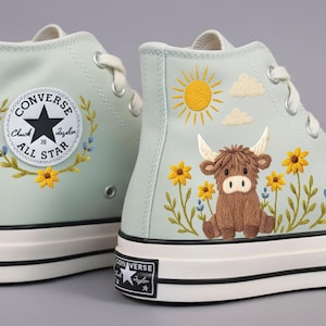 Puede incluir: Zapatillas altas verde claro con detalles bordados. Una zapatilla tiene el logo de Converse, la otra una vaca de las Tierras Altas marrón, flores amarillas, un sol y nubes. Cordones y suelas blancas.