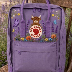 May include: A purple backpack featuring an embroidered highland cow, lavender sprigs, butterflies, and colorful wildflowers. The front displays a circular Fjällräven Kånken logo patch, two top handles, a zippered main compartment, and a front pocket.