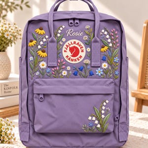 以下が含まれることがあります： ラベンダー色のバックパックで、刺繍された野花と白い筆記体で書かれた「Rosie」という名前が特徴です。前面にはFjällräven Kånkenの円形ロゴパッチ、ファスナー付きポケット、2つのトップハンドルが付いています。花のデザインには黄色、白、青の花が含まれています。