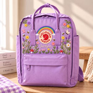 May include: A light purple backpack featuring an embroidered design of a rainbow, flowers, mushrooms, bees, and a butterfly. The circular Fjällräven Kånken logo is centered above the name Jazzlynn, with two top handles and a front zippered pocket.