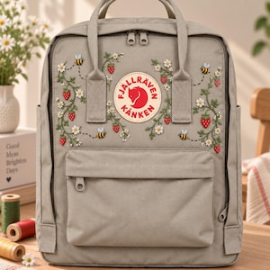 May include: A light gray backpack featuring an embroidered pattern of red strawberries, white daisies, and yellow bees. The front displays a circular Fjällräven Kånken logo patch in red and cream. It includes a top handle, dual zippers, and a front pocket.