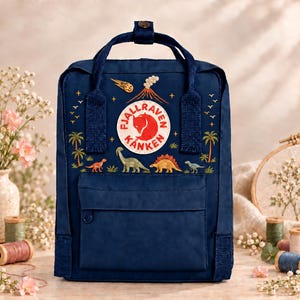 Peut inclure: Un sac à dos bleu marine avec un écusson circulaire Fjällräven Kånken, brodé d'une éruption volcanique, d'une comète souriante, de palmiers et de divers dinosaures colorés, dont un sauropode au long cou et un stégosaure à épines.