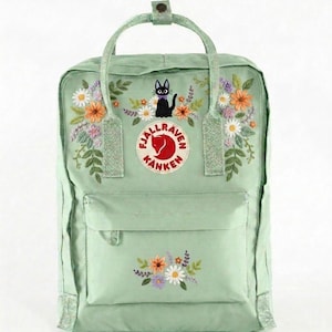 Könnte beinhalten: Ein hellgrüner Rucksack mit einer Stickerei eines schwarzen Katers mit violetter Fliege, umgeben von bunten Blumenarrangements. Das kreisförmige Fjällräven Kånken-Logo ist mittig auf der Vorderseite platziert, ergänzt durch florale Stickereien.