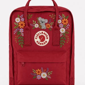 May include: A deep red backpack featuring a circular Fjällräven Kånken logo patch on the front. The bag is decorated with embroidered floral patterns and a small gray koala illustration. It includes a front zippered pocket and top handles.