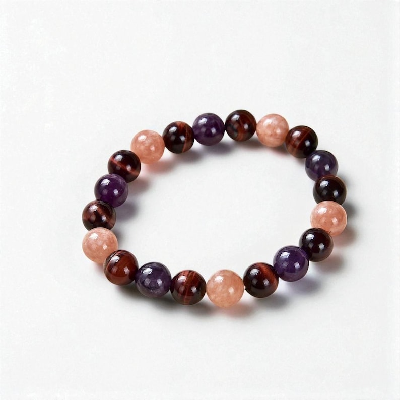 May include: A beaded bracelet featuring a variety of colorful gemstones. The bracelet includes round beads in shades of purple, red-brown, and peach. The beads are strung together to form a complete circle.