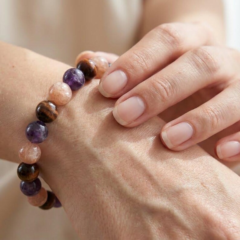 May include: A beaded bracelet featuring a mix of natural gemstones. The bracelet includes round beads in shades of purple, brown, and peach. The bracelet is worn on a wrist, with a hand gently touching it.