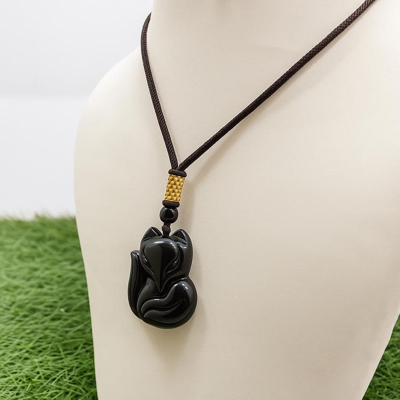 May include: A black fox pendant necklace. The pendant is a glossy, carved fox figure suspended from a brown cord. The cord is attached to a gold-colored bead and a black bead. The necklace is displayed on a white mannequin.