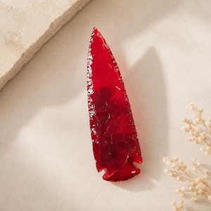 May include: A translucent, deep red arrowhead with a sharp, pointed tip and a notched base. The surface features a textured, flaked appearance characteristic of knapped stone, creating a glossy finish that catches light across its elongated form.
