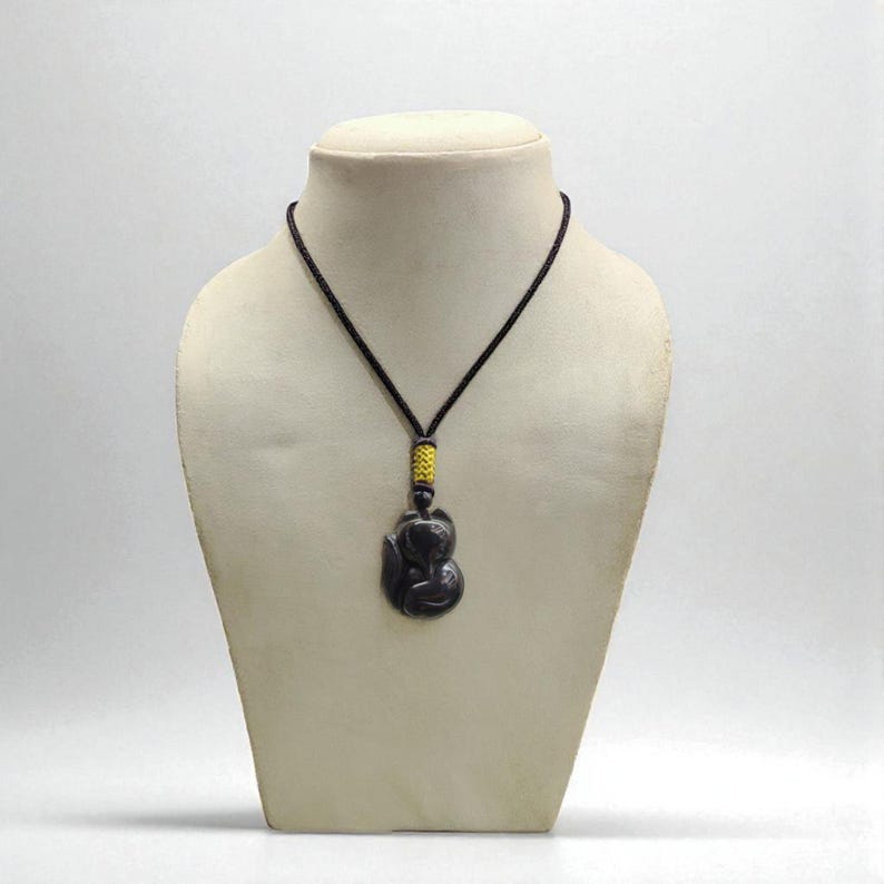 May include: A dark gray pendant necklace with a black cord. The pendant is a stylized figure, and the cord has a yellow and black accent. The necklace is displayed on a mannequin.