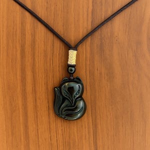 May include: A black fox pendant necklace. The pendant is a glossy, carved fox figure. The necklace cord is dark brown, with a cream-colored accent near the top. The background is a wooden surface.