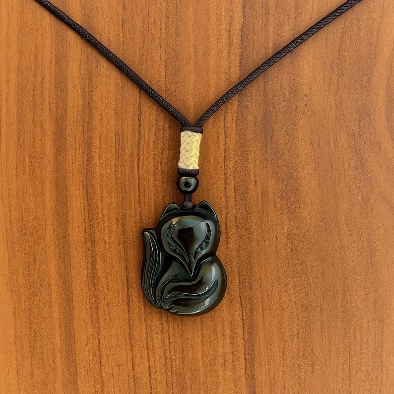 May include: A black fox pendant necklace. The pendant is a glossy, carved fox figure. The necklace cord is dark brown, with a cream-colored accent near the top. The background is a wooden surface.