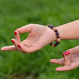 May include: A multi-colored beaded bracelet with purple, brown, and peach-colored beads. The bracelet is on a wrist with red nail polish. The hand is in a yoga mudra position.