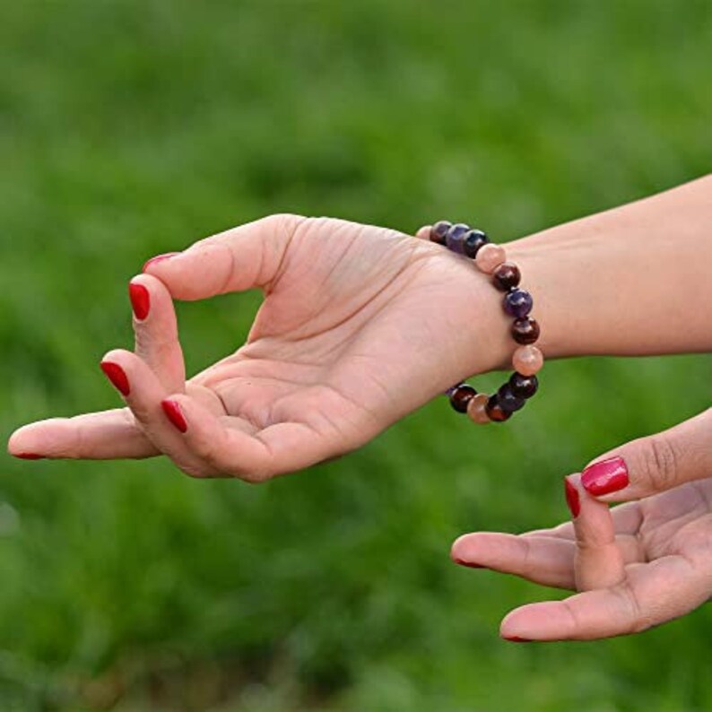 May include: A multi-colored beaded bracelet with purple, brown, and peach-colored beads. The bracelet is on a wrist with red nail polish. The hand is in a yoga mudra position.