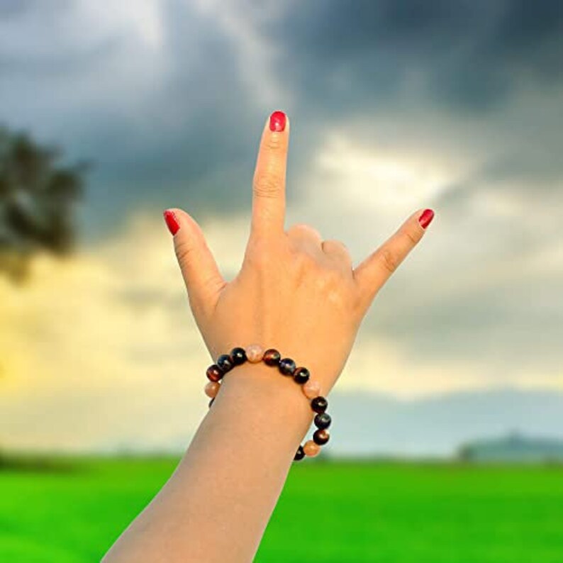 May include: A beaded bracelet with brown, black, and tan stones is worn on a wrist. The hand is making the "I love you" sign with red nail polish. The background is a blurred green field and cloudy sky.