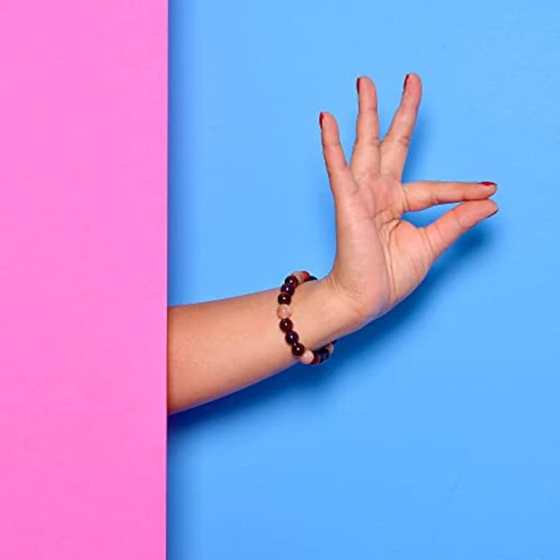May include: A beaded bracelet with dark brown and pinkish-tan stones is worn on a wrist. The background is divided into two vertical sections: a bright pink on the left and a solid blue on the right. The hand is making a gesture.