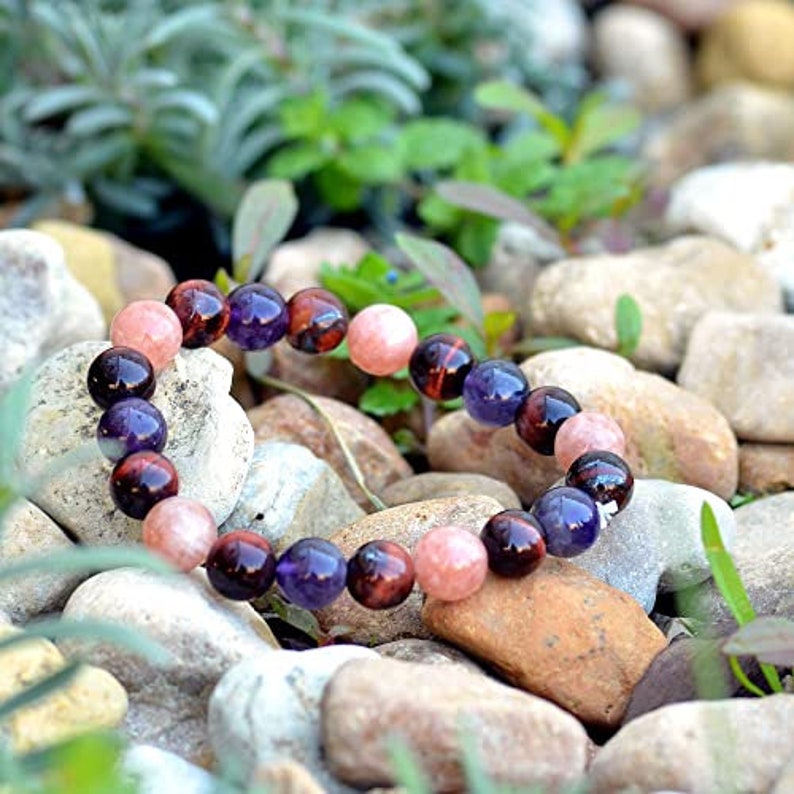 May include: A beaded bracelet featuring a mix of round gemstones in shades of pink, purple, and brown. The bracelet is displayed on a bed of small rocks and greenery, showcasing its natural colors and textures.