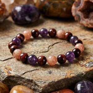 May include: A beaded bracelet featuring a mix of gemstones. The bracelet includes round beads in shades of purple, red-brown, and peach. The bracelet is displayed on a textured, neutral-toned surface, with other gemstones in the background.