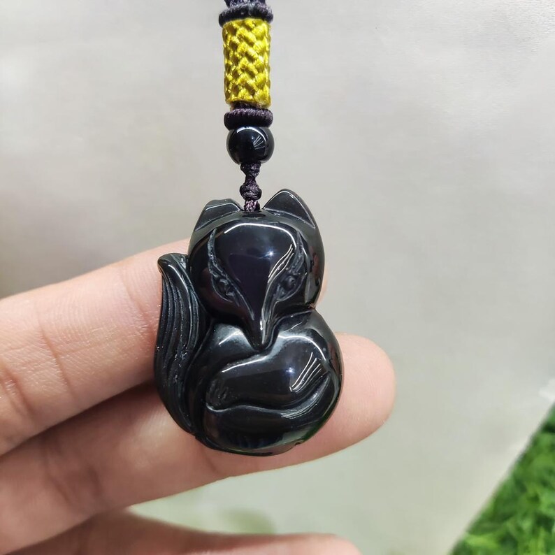 May include: A black fox pendant carved from a glossy material. The fox is detailed with carved features and is suspended from a yellow and black cord necklace. The pendant is held up by a person's fingers.
