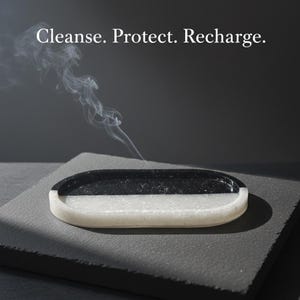 Pode incluir: Um porta-incenso oval, dividido em seções pretas e brancas, com volutas de fumaça subindo. O texto "Cleanse. Protect. Recharge." é exibido acima do porta-incenso. Ele está sobre uma superfície cinza escura.